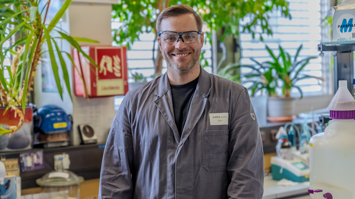 Thomas Nack lächelt in die Kamera. Er trägt eine Schutzbrille und einen grauen Laborkittel. Im Hintergrund ist ein Labor zu sehen.