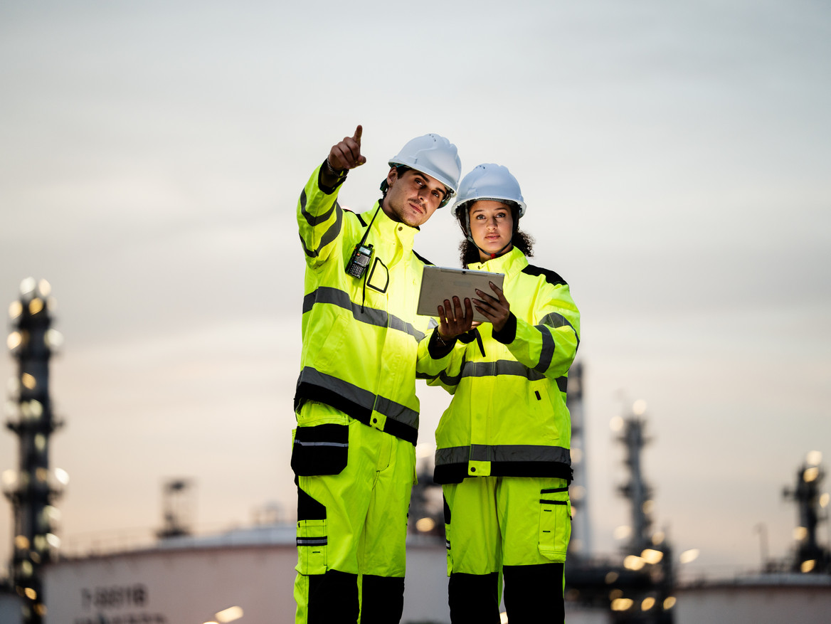 Zwei Chemiebeschäftigte stehen auf der Außenanlage ihres Betriebs und richten vor dämmerigem Hintergrund einen prüfenden Blick auf die Anlagen.
