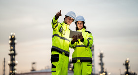 Zwei Chemiebeschäftigte stehen auf der Außenanlage ihres Betriebs und richten vor dämmerigem Hintergrund einen prüfenden Blick auf die Anlagen.