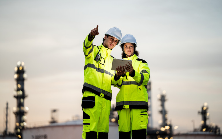Zwei Chemiebeschäftigte stehen auf der Außenanlage ihres Betriebs und richten vor dämmerigem Hintergrund einen prüfenden Blick auf die Anlagen.