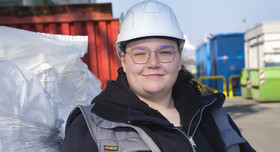 Eine junge Frau mit Schutzhelm steht vor großen Containern.