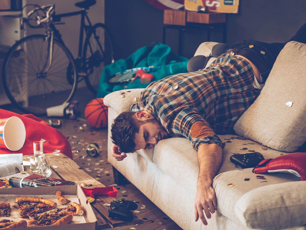 Junger Mann hängt nach einer Party kopfüber auf seinem Sofa.