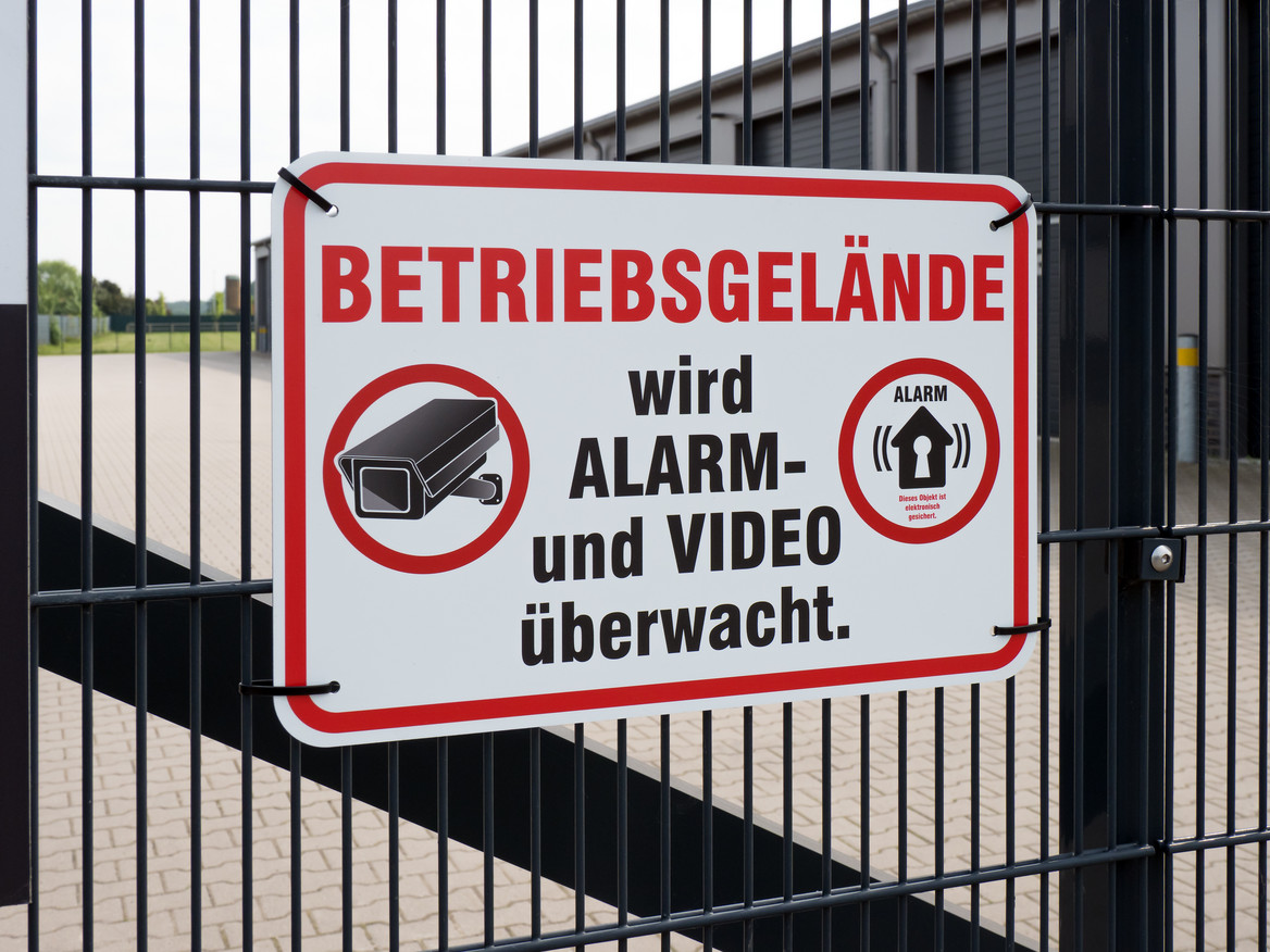 An einem Fabrikzaun hängt ein Schild: "Betriebsgelände wird Alarm- und Video überwacht." 