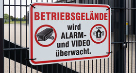 An einem Fabrikzaun hängt ein Schild: "Betriebsgelände wird Alarm- und Video überwacht." 