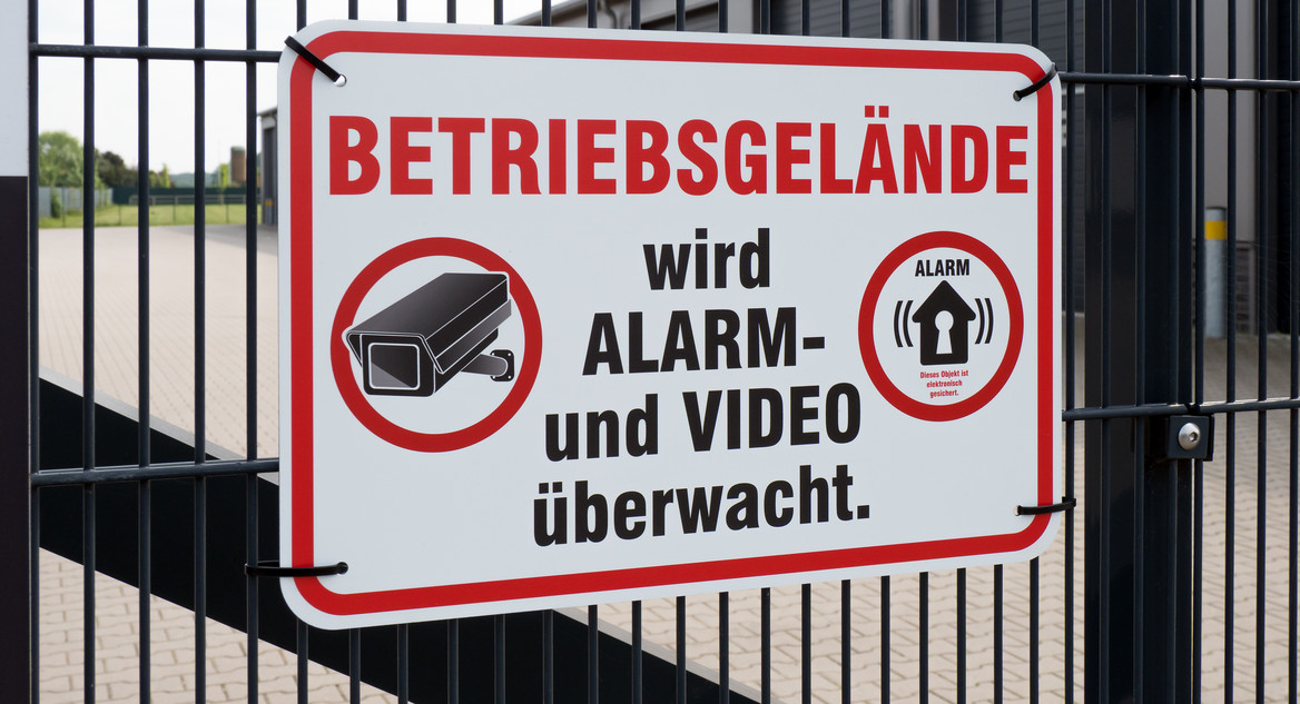 An einem Fabrikzaun hängt ein Schild: "Betriebsgelände wird Alarm- und Video überwacht." 