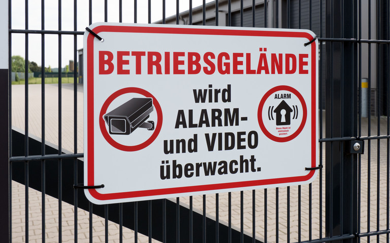 An einem Fabrikzaun hängt ein Schild: "Betriebsgelände wird Alarm- und Video überwacht." 