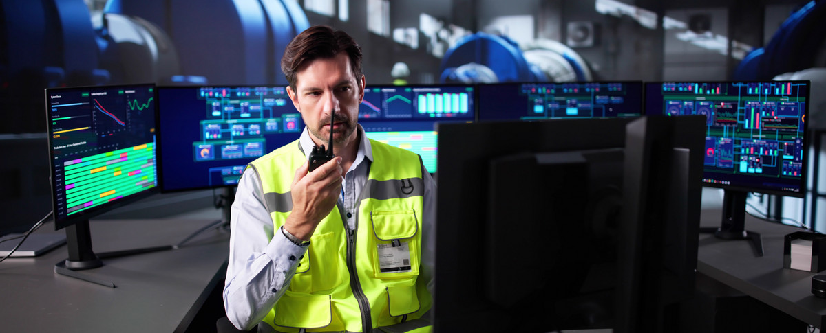 Ingenieur mit Funkgerät in der Leitzentrale eines Produktonsbetriebes: Er spricht und blickt konzentriert auf die Monitore.