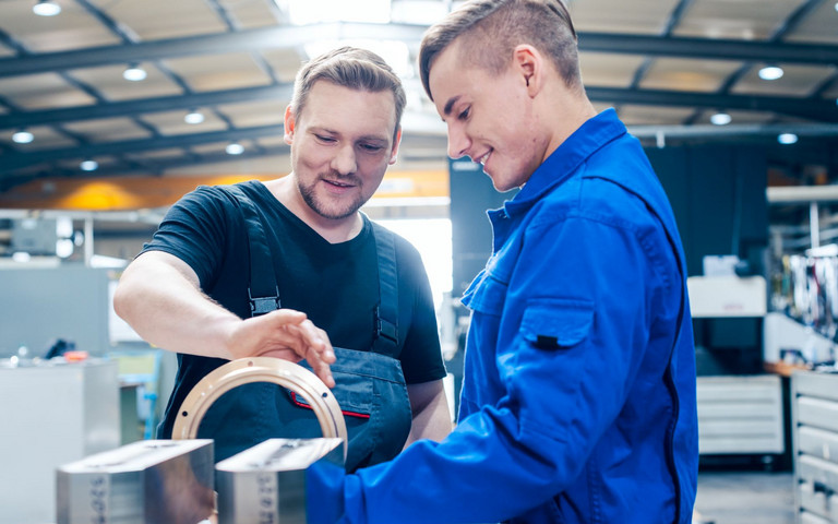 Ein freundlicher Meister und ein interessierter Azubi im Blaumann begutachten in der Werkstatt ein Arbeitsergebnis.