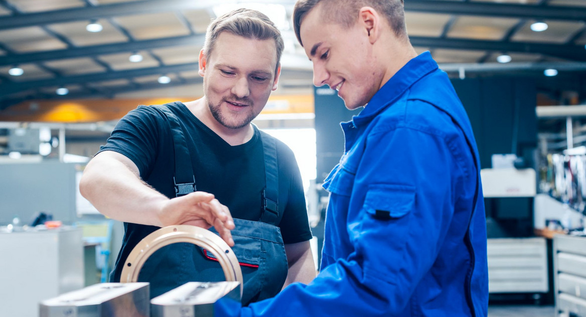 Ein freundlicher Meister und ein interessierter Azubi im Blaumann begutachten in der Werkstatt ein Arbeitsergebnis.