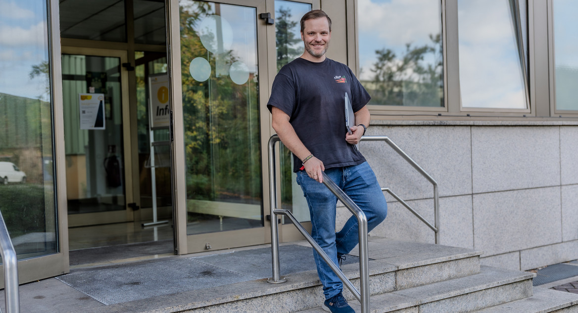 Thomas Nack steht lächelnd auf einer Treppe vor dem Eingang eines Bürogebäudes mit großer Glastür. Er trägt ein dunkles T-Shirt, Jeans und Sneaker und hält einen Laptop unter dem Arm.