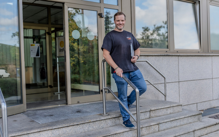 Thomas Nack steht lächelnd auf einer Treppe vor dem Eingang eines Bürogebäudes mit großer Glastür. Er trägt ein dunkles T-Shirt, Jeans und Sneaker und hält einen Laptop unter dem Arm.