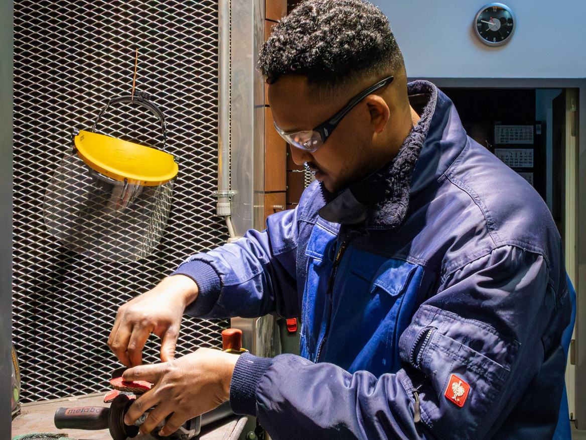 Regan Kifle Negash arbeitet mit Schutzbrille und Arbeitsjacke an einer Maschine.
