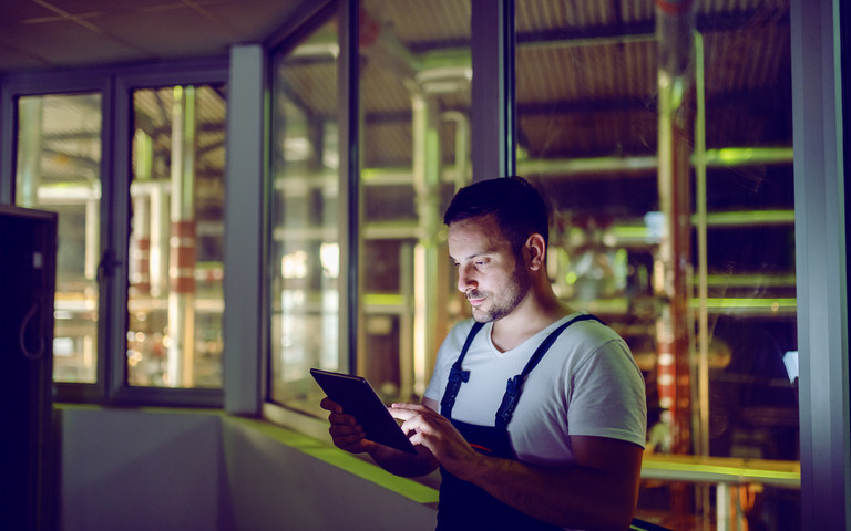 Ein Mann mit Tablet und blauer Latzhose steht in einer wenig beleuchteten Fabrik.