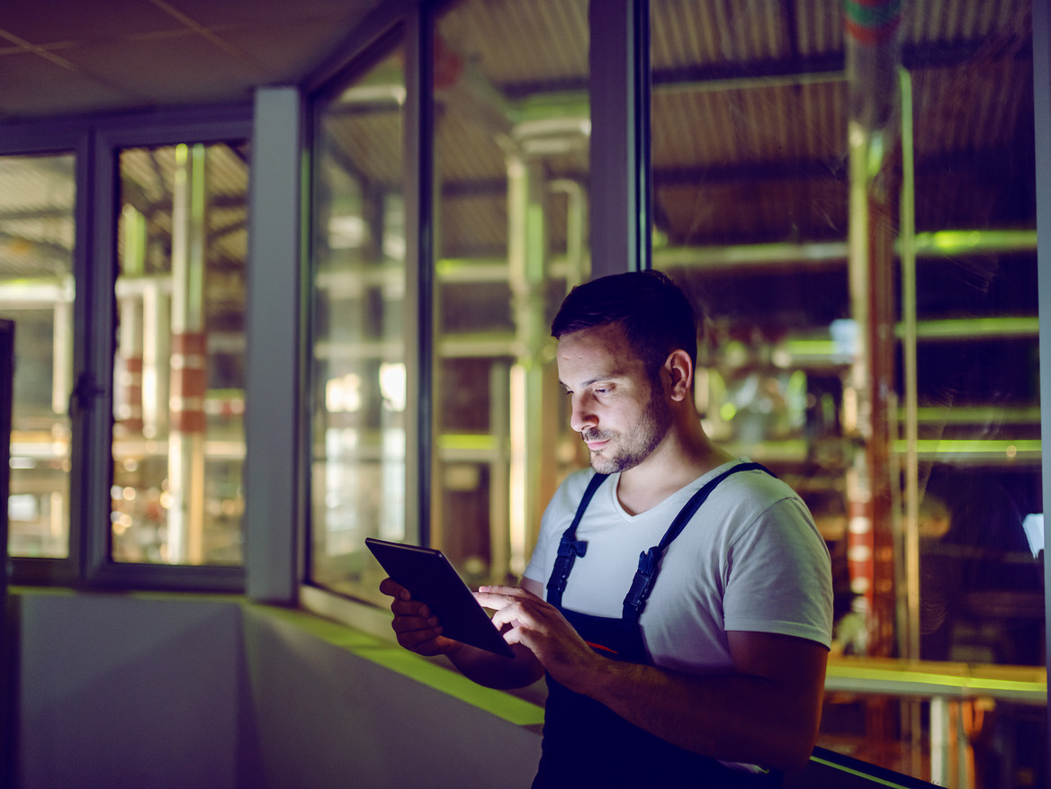 Ein Mann mit Tablet und blauer Latzhose steht in einer wenig beleuchteten Fabrik.