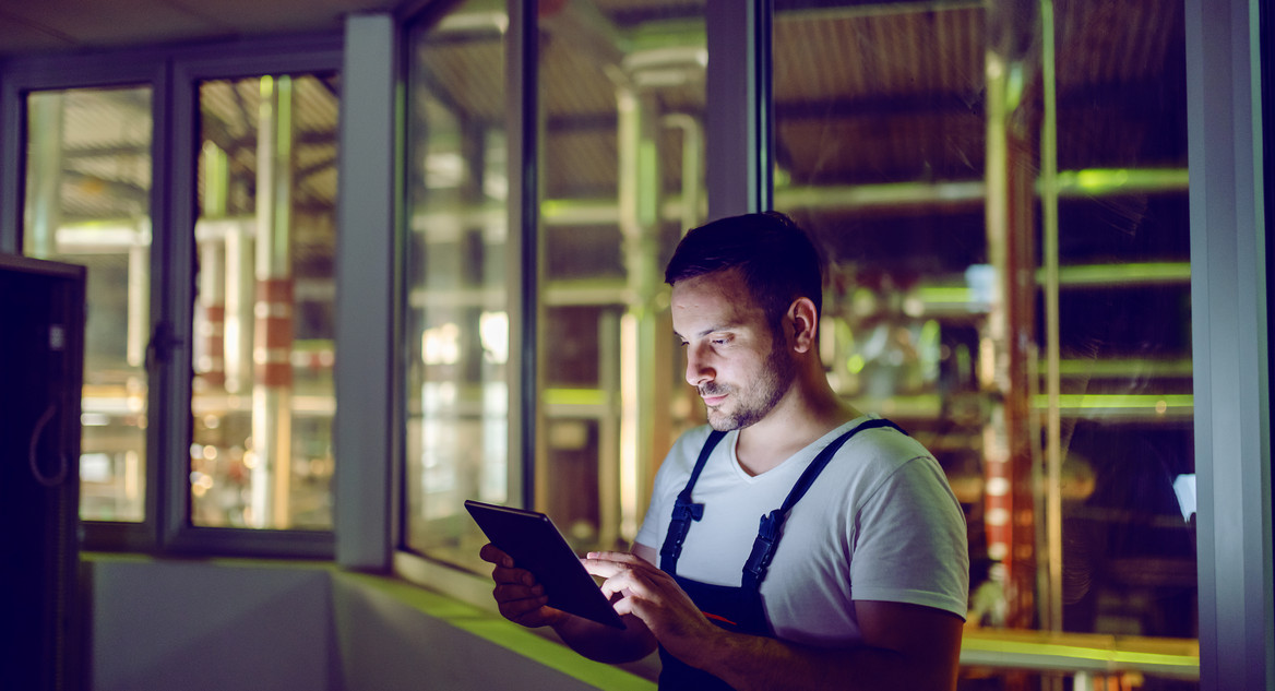 Ein Mann mit Tablet und blauer Latzhose steht in einer wenig beleuchteten Fabrik.