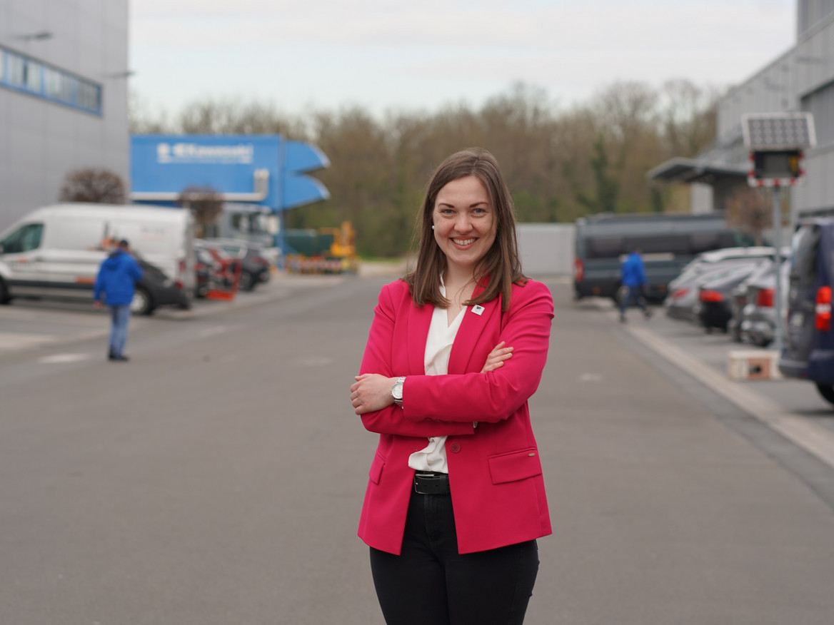 Clara Scherner steht auf dem Werksgelände, trägt Businesskleidung und schaut lächelnd in die Kamera.