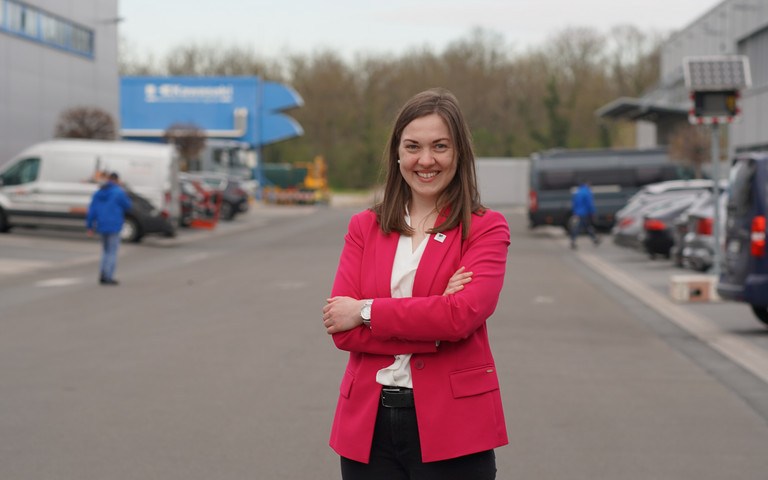 Clara Scherner steht auf dem Werksgelände, trägt Businesskleidung und schaut lächelnd in die Kamera.