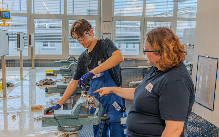 Ausbilderin Kristina Eckert zeigt einem Auszubildenden an einer Werkbank einen Arbeitsschritt. Er bedient eine Maschine, während sie auf das Werkstück weist.