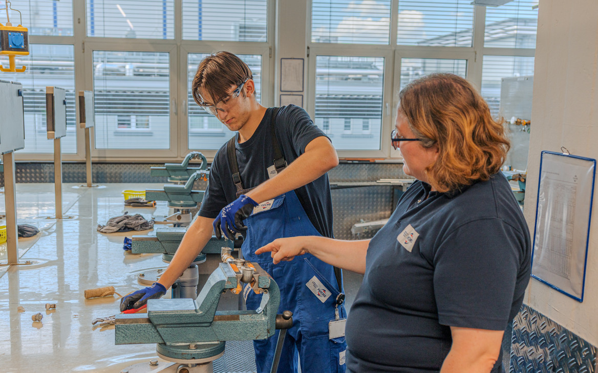 Ausbilderin Kristina Eckert zeigt einem Auszubildenden an einer Werkbank einen Arbeitsschritt. Er bedient eine Maschine, während sie auf das Werkstück weist.