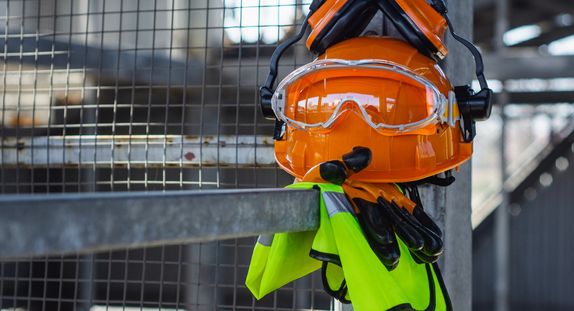 Persönliche Schutzausrüstung mit Helm, Gehörschutz, Brille, Handschuhen und Sicherheitsweste ist auf einer Metallabsperrung abgelegt.