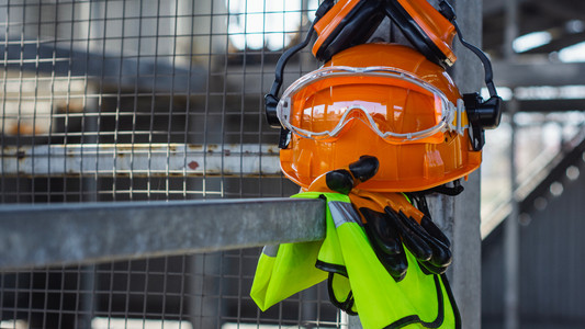 Persönliche Schutzausrüstung mit Helm, Handschuhen, Weste und Schutzbrille auf einem Werkzaun.