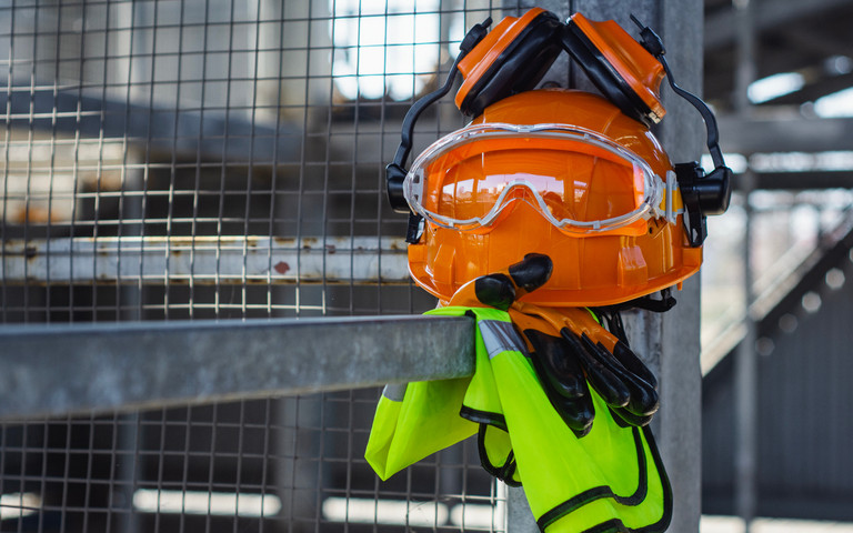 Persönliche Schutzausrüstung mit Helm, Gehörschutz, Brille, Handschuhen und Sicherheitsweste ist auf einer Metallabsperrung abgelegt.