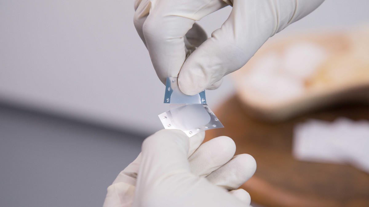 Eine Fachkraft hält ein winziges Pflaster der Firma LTS jeweils zwischen Daumen und Zeigefingern der handschuhgeschützten Hände. Diese Pflaster geben über die Haut Wirkstoffe an die Patientinnen und Patienten ab.