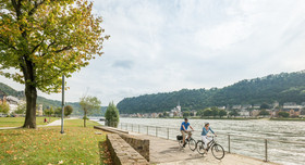 Zwei Radfahrer fahren an einem großen Fluss entlang.