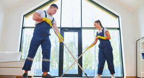 Junges Paar in blauen Overalls mit Wischmopp beim Hausputz in sonnendurchflutetem Raum.