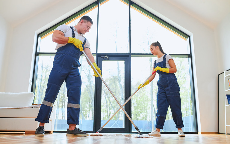 Junges Paar in blauen Overalls mit Wischmopp beim Hausputz in sonnendurchflutetem Raum.