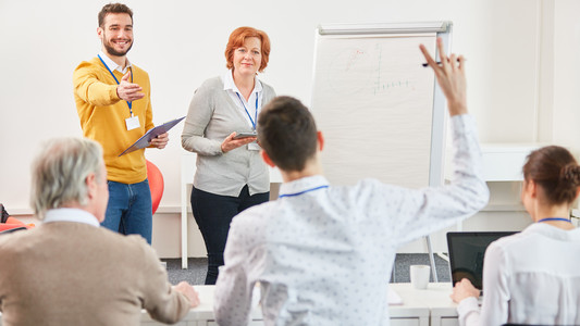 Ein freundliches Dozenten-Duo steht vor einem Flipchart und wendet sich aktiven Weiterbildungsteilnehmenden zu.