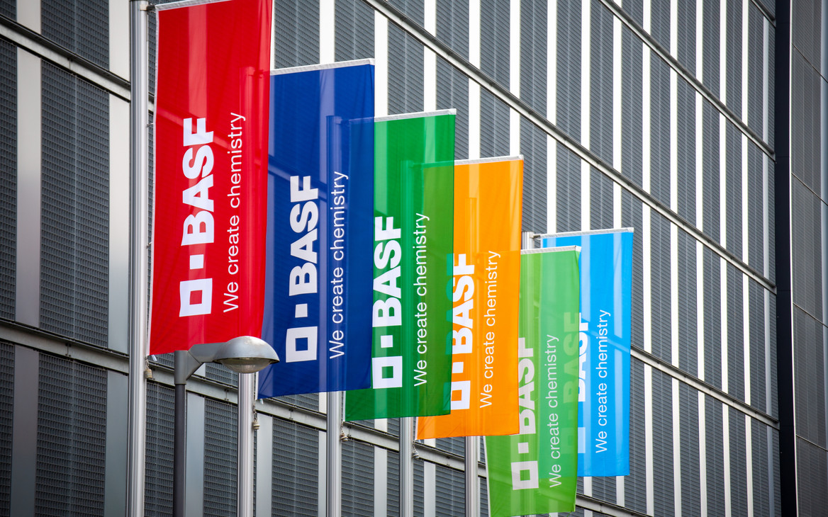 Mehrere bunte BASF-Fahnen in Rot, Blau, Grün und Orange mit der Aufschrift „BASF – We create chemistry“ hängen an Fahnenmasten vor dem Firmengebäude in Ludwigshafen.