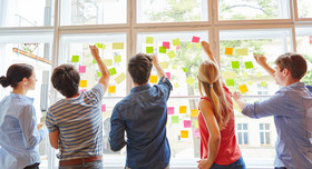 Ein fünfköpfiges Workshopteam heftet Post-its an eine sonnendurchstrahlte Fensterfront.