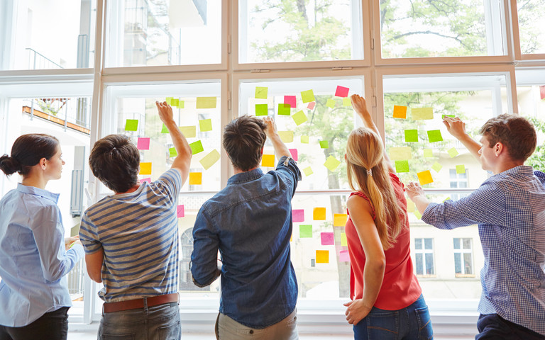 Ein fünfköpfiges Workshopteam heftet Post-its an eine sonnendurchstrahlte Fensterfront.
