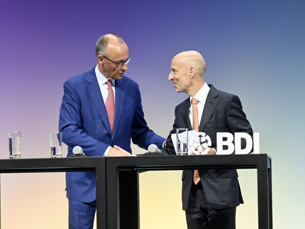 Bundeskanzler Friedrich Merz und BDI-Präsident Peter Leibinger stehen an einem Tisch und sprechen miteinander. 