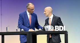 Bundeskanzler Friedrich Merz und BDI-Präsident Peter Leibinger stehen an einem Tisch und sprechen miteinander. 