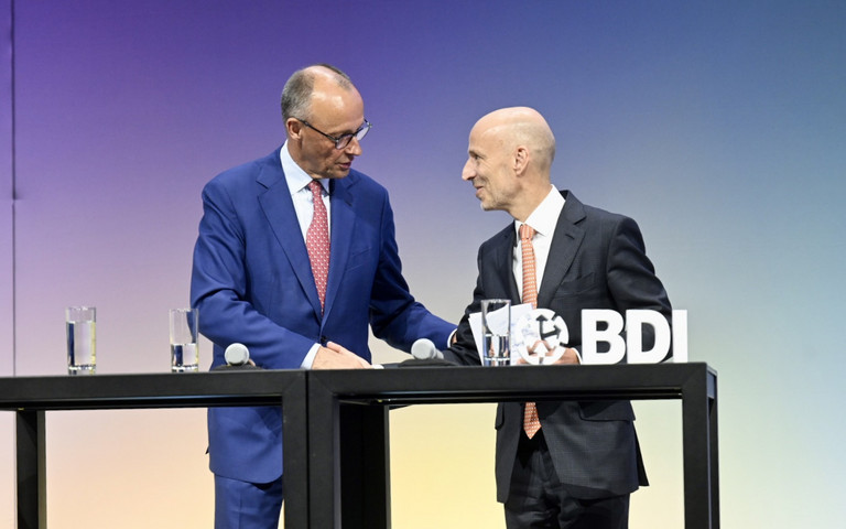 Bundeskanzler Friedrich Merz und BDI-Präsident Peter Leibinger stehen an einem Tisch und sprechen miteinander. 