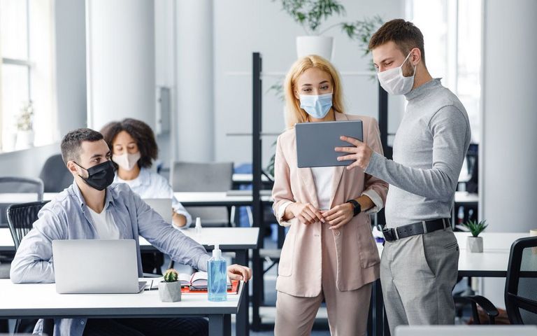 Teamwork in corporate company and social distance during coronavirus epidemic. Male manager in protective mask shows tablet to workers in interior.