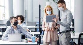 Teamwork in corporate company and social distance during coronavirus epidemic. Male manager in protective mask shows tablet to workers in interior.