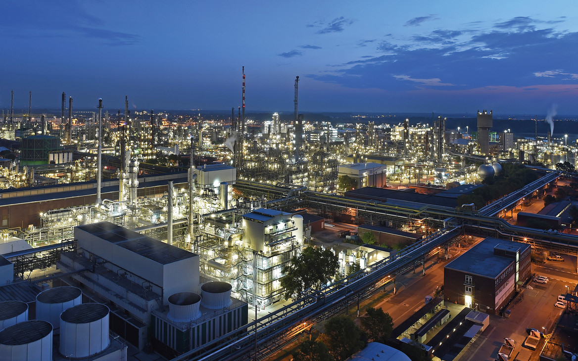 Nächtliche Luftaufnahme des BASF-Verbundstandorts Ludwigshafen: Das weitläufige Industrieareal erstreckt sich bis zum Horizont und ist hell erleuchtet. Zu sehen sind zahlreiche Produktionsanlagen, Türme, Rohrleitungen und Gebäude, durchzogen von beleuchteten Straßen. Im Hintergrund ist der Abendhimmel in Blau- und Rottönen zu erkennen.