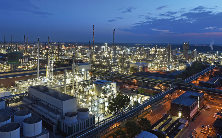 Nächtliche Luftaufnahme des BASF-Verbundstandorts Ludwigshafen: Das weitläufige Industrieareal erstreckt sich bis zum Horizont und ist hell erleuchtet. Zu sehen sind zahlreiche Produktionsanlagen, Türme, Rohrleitungen und Gebäude, durchzogen von beleuchteten Straßen. Im Hintergrund ist der Abendhimmel in Blau- und Rottönen zu erkennen.