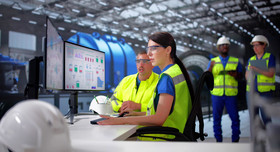 Ein Ingenieursteam mit Sicherheitswesten am Leitstand. Sie steuern per Monitor industrielle Prozesse.