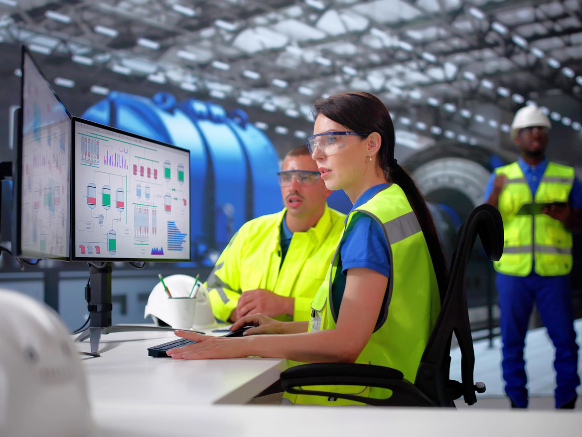 Ein Ingenieursteam mit Sicherheitswesten am Leitstand. Sie steuern per Monitor industrielle Prozesse.