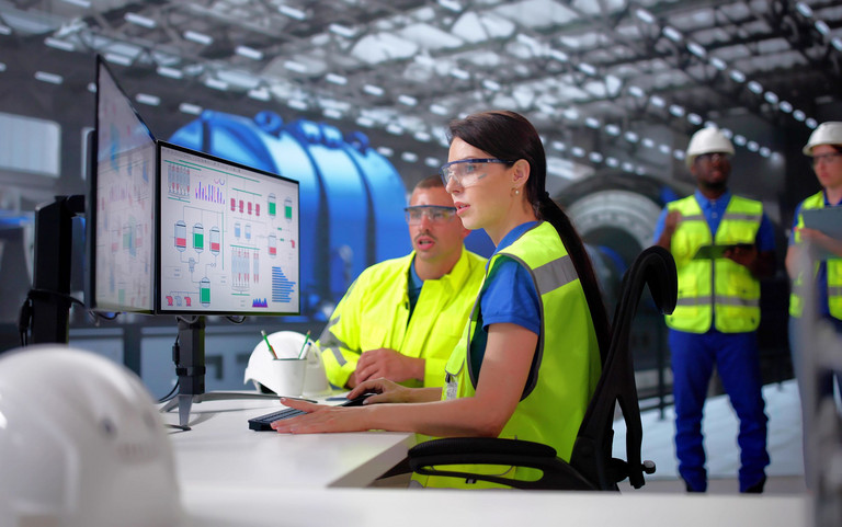 Ein Ingenieursteam mit Sicherheitswesten am Leitstand. Sie steuern per Monitor industrielle Prozesse.