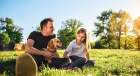 Vater, Tochter und Hund entspannen bei sonnigem Wetter auf einer Parkwiese.