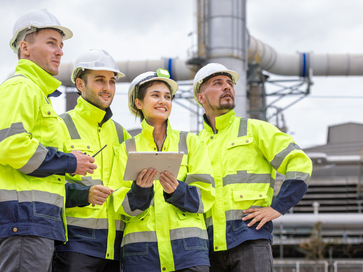 Ein vierköpfiges Fachkräfteteam in Sicherheitskleidung steht außen an einer Chemieanlage und blickt positiv-beobachtend in dieselbe Richtung.