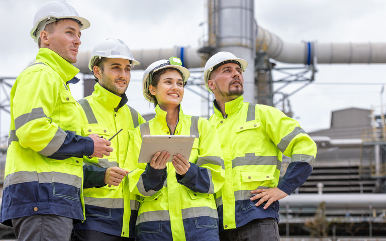 Ein vierköpfiges Fachkräfteteam in Sicherheitskleidung steht außen an einer Chemieanlage und blickt positiv-beobachtend in dieselbe Richtung.