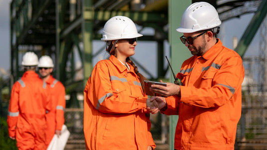 Zwei Chemie-Führungskräfte beim kollegialen Handshake auf dem Außengelände einer Chemieanlage. Zwei weitere Kollegen stehen mit einem Anlagenplan im Hintergrund.