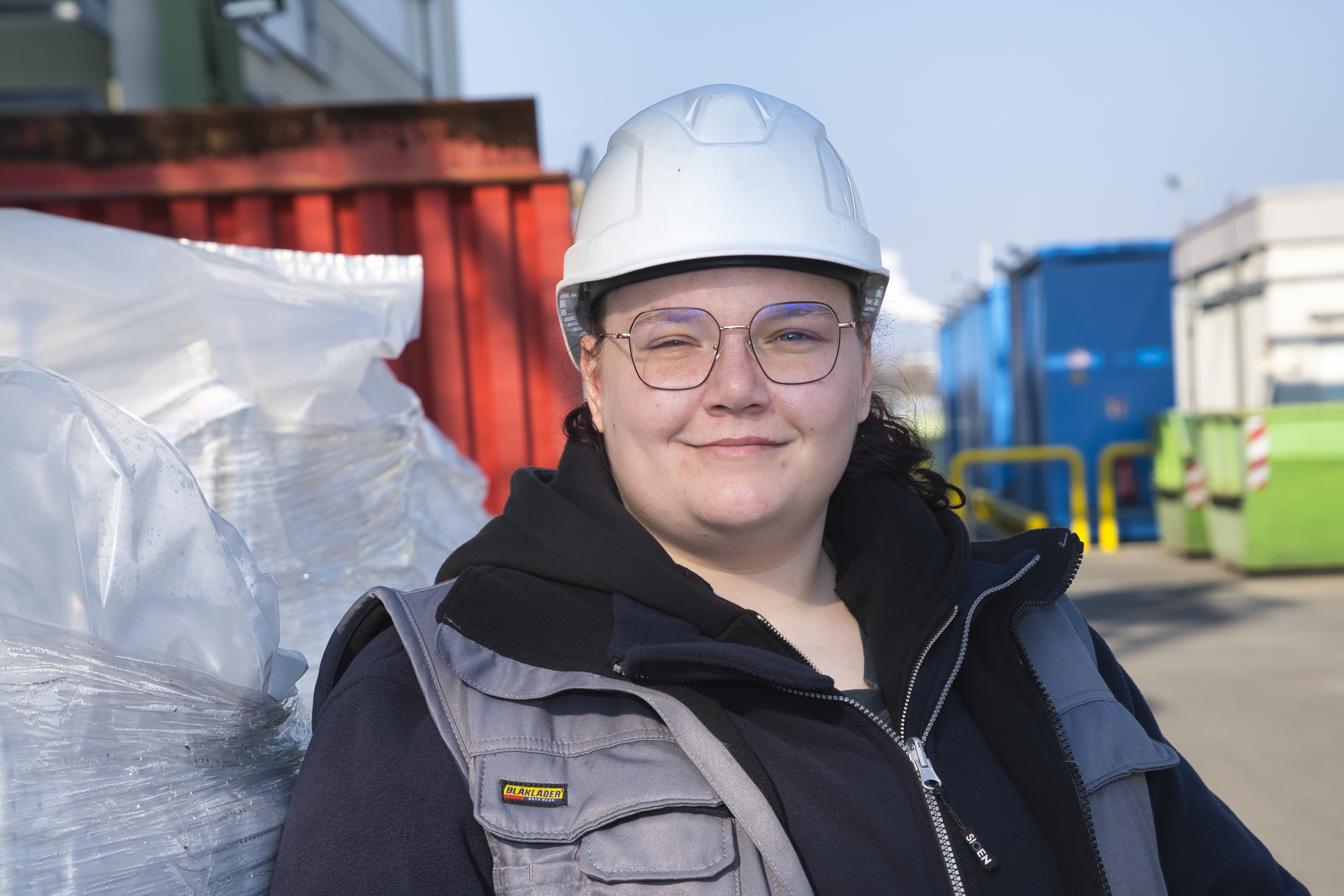 Eine junge Frau mit Schutzhelm steht vor großen Containern.